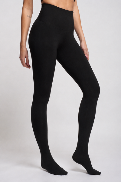 A person wearing TLC Sport Extra Strong Compression Tummy Control Leggings with Feet in opaque black, standing against a plain light background. Only the lower body and part of the left arm are visible.