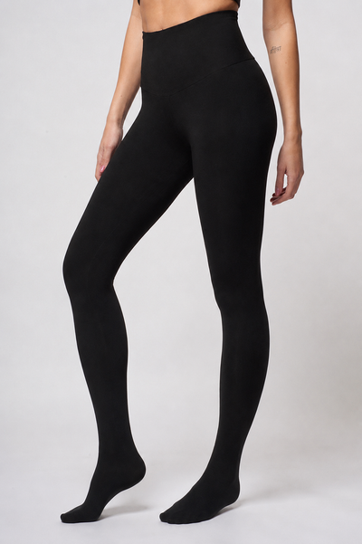 A person wearing TLC Sport Lightweight Strong Compression Tummy Control Leggings with Feet stands barefoot against a plain light background, arms relaxed by their sides.
