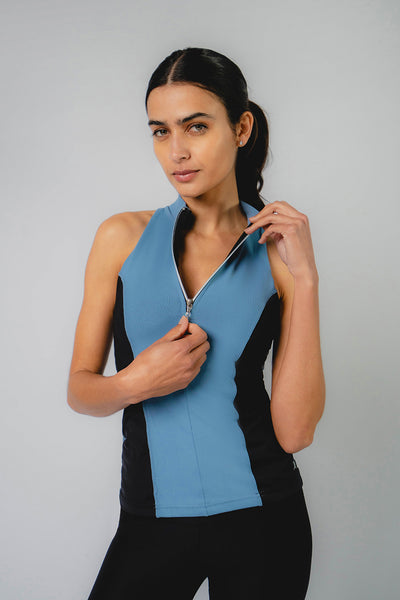 A woman with dark hair in a ponytail wears the TLC Sport Zip Front Polo Top in Black/Airforce Blue, a sleeveless workout top with an adjustable front zip partially unzipped at the collar, standing against a plain light background.